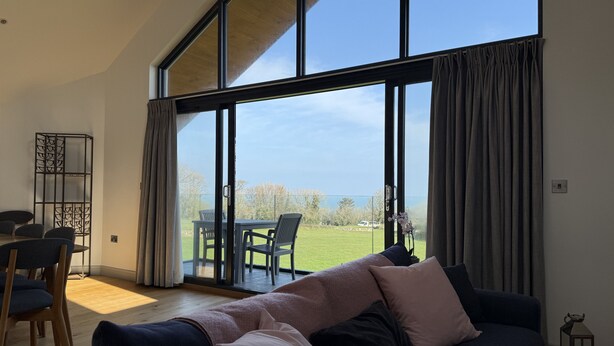 A modern living room with a large window, a sofa, a dining table with chairs, and a view of a green landscape. - Fishers View - a lodge that sleeps 10 guests in 5 bedrooms (St Ives)