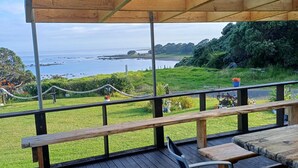 Una terraza de madera con bancos y una mesa, con vistas a un área verde y el océano.