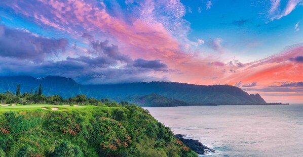 A coastal landscape with a golf course, mountains, and a sunset.