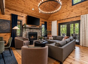 A cozy living room with a stone fireplace, comfortable seating, a flat-screen TV, and large windows offering a view of the outdoors.