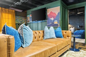 A brown leather sofa with blue and white cushions, a flat-screen TV mounted on the wall, and a poster depicting a scenic route.