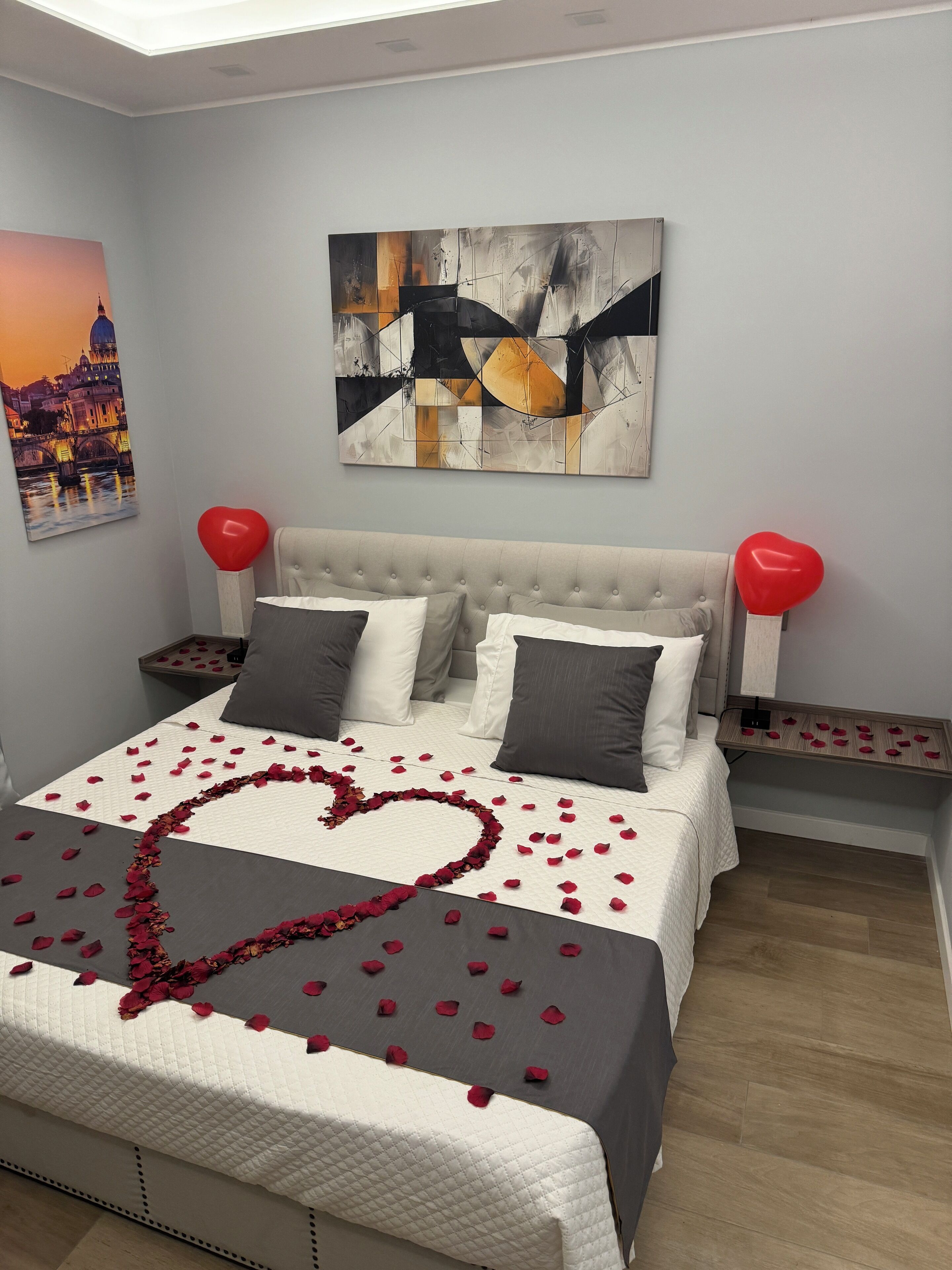 Une chambre à coucher avec un lit, une composition en forme de cœur faite de pétales de roses, un tableau représentant un paysage urbain et une œuvre d’art abstrait accrochée au mur.