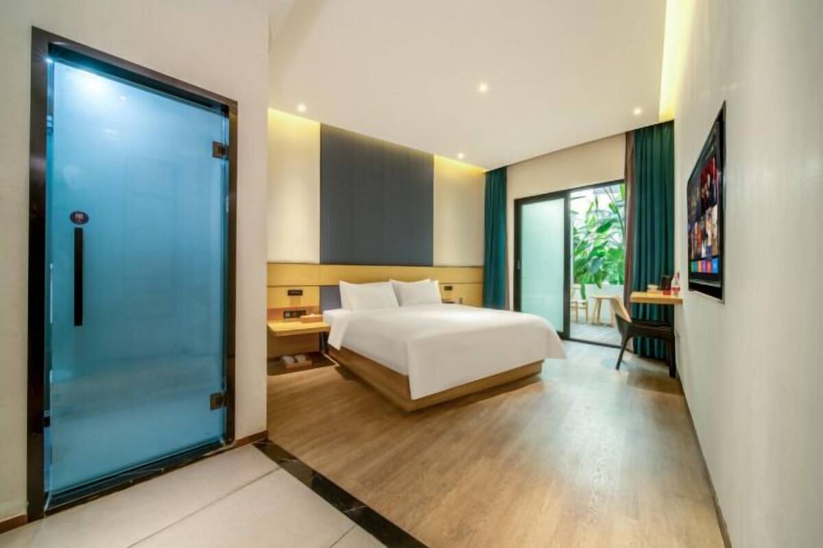 Habitación de hotel con cama, escritorio, silla, puerta corrediza y vistas a la vegetación exterior.
