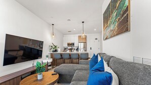 Uma sala de estar moderna com uma grande televisão de tela plana, uma mesa de centro redonda de madeira e um sofá modular cinza com almofadas azuis e brancas.