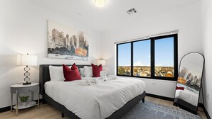 Um quarto moderno com uma cama grande, uma pintura de paisagem urbana, uma janela com vista e uma mesa de cabeceira com abajur.