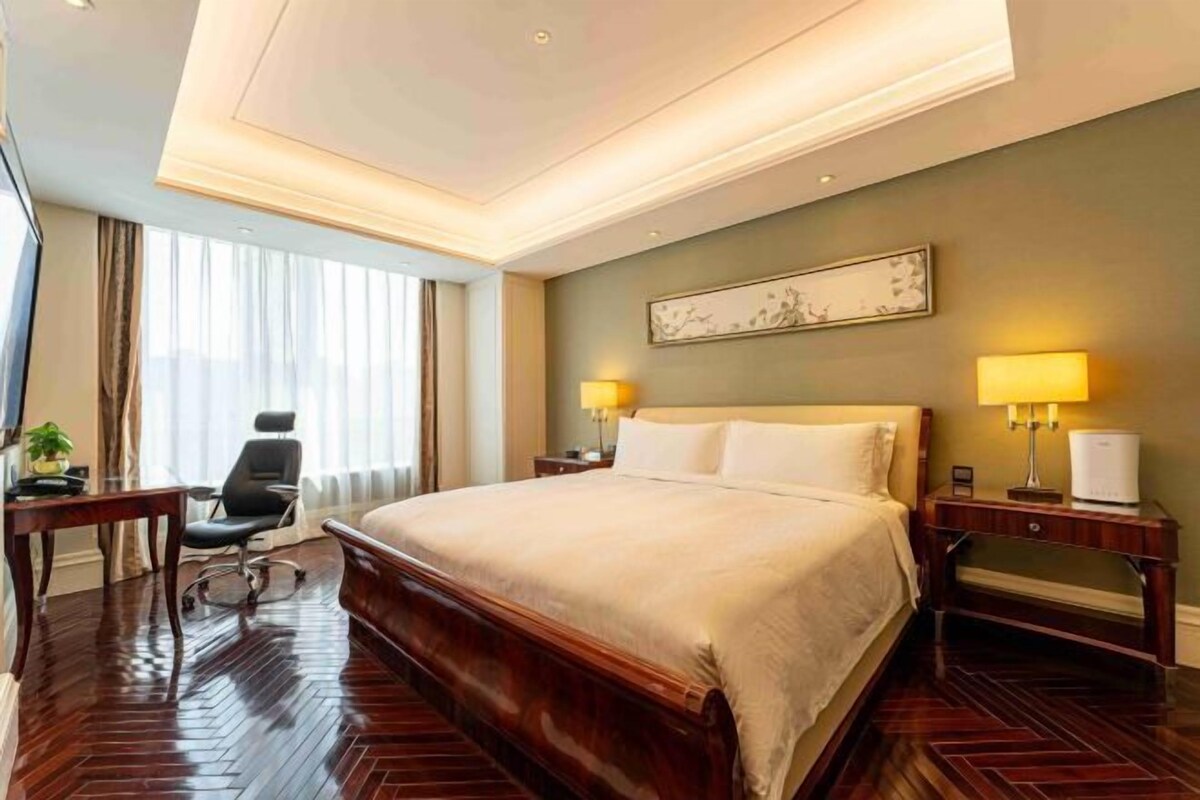 Habitación de hotel con una cama grande, dos mesitas de noche con lámparas, un escritorio con silla y una ventana con cortinas.