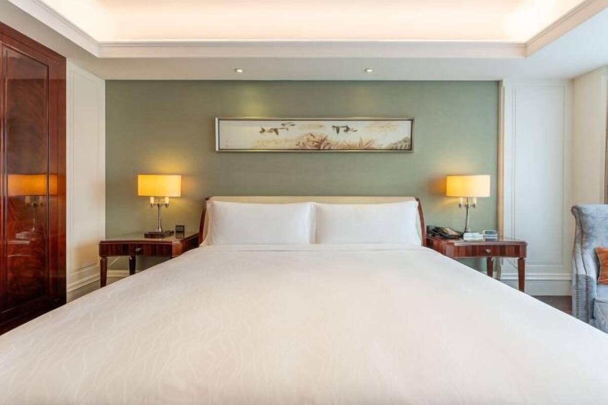 A hotel room with a large bed, two bedside tables with lamps, a painting on the wall, and a wooden wardrobe.