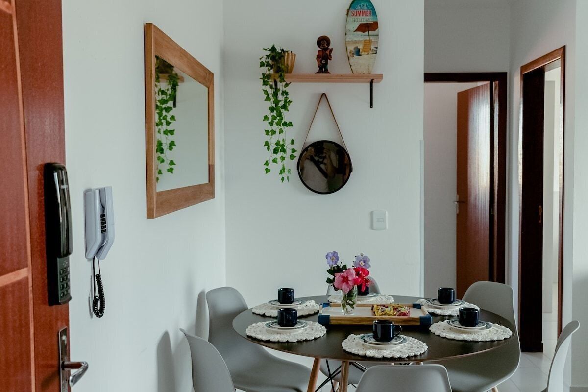 Un comedor con una mesa redonda dispuesta para cuatro personas, rodeada de sillas. Hay un espejo en la pared, un teléfono y una estantería con objetos decorativos.