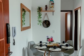 A dining area with a round table set for four, surrounded by chairs. There is a mirror on the wall, a telephone, and a shelf with decorative items.