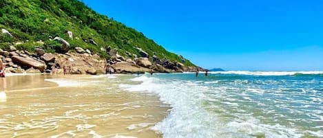 A beach with clear waters, a rocky shoreline, and a lush green hill in the background.