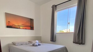 Een net opgemaakt bed met een grijze deken en kussens, een groot raam met gordijnen en een schilderij van een zonsondergang aan de muur.