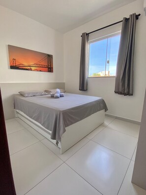 A neatly made bed with a gray bedspread and pillows, a large window with curtains, and a sunset painting on the wall.