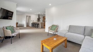 A modern living room with a grey sofa, two wicker chairs with cushions, a wooden coffee table, and a kitchen area in the background. - Calm at Leitrum - Cromwell Holiday Home (Cromwell)