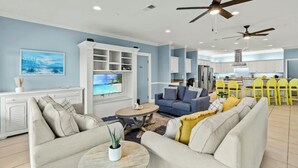 A spacious living room with a large TV, sofas, and a coffee table. There are ceiling fans and a kitchen in the background. - Rates Reduced-Canary-Oceanfront-Shared Pool-Game Room-4000 sqft (North Myrtle Beach)