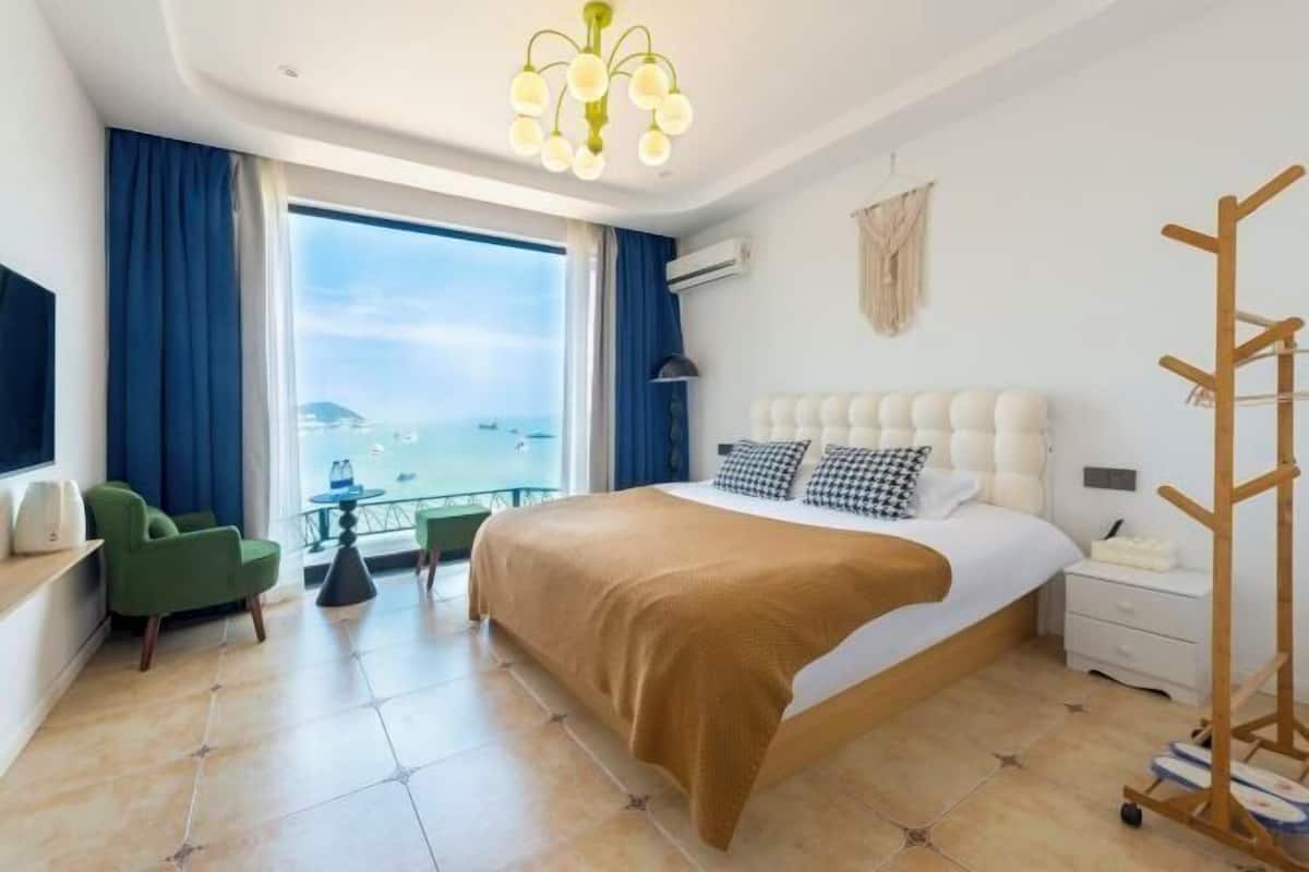 Ein Hotelzimmer mit einem großen Bett, einem grünen Sessel, einem kleinen Tisch, einem Balkon mit Aussicht und einem Holz-Kleiderständer.