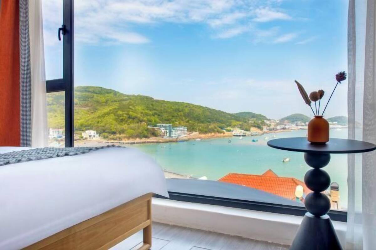 Ein Schlafzimmer mit großem Fenster und Blick aufs Meer, einem Bett und einem kleinen Tisch mit einer Vase.