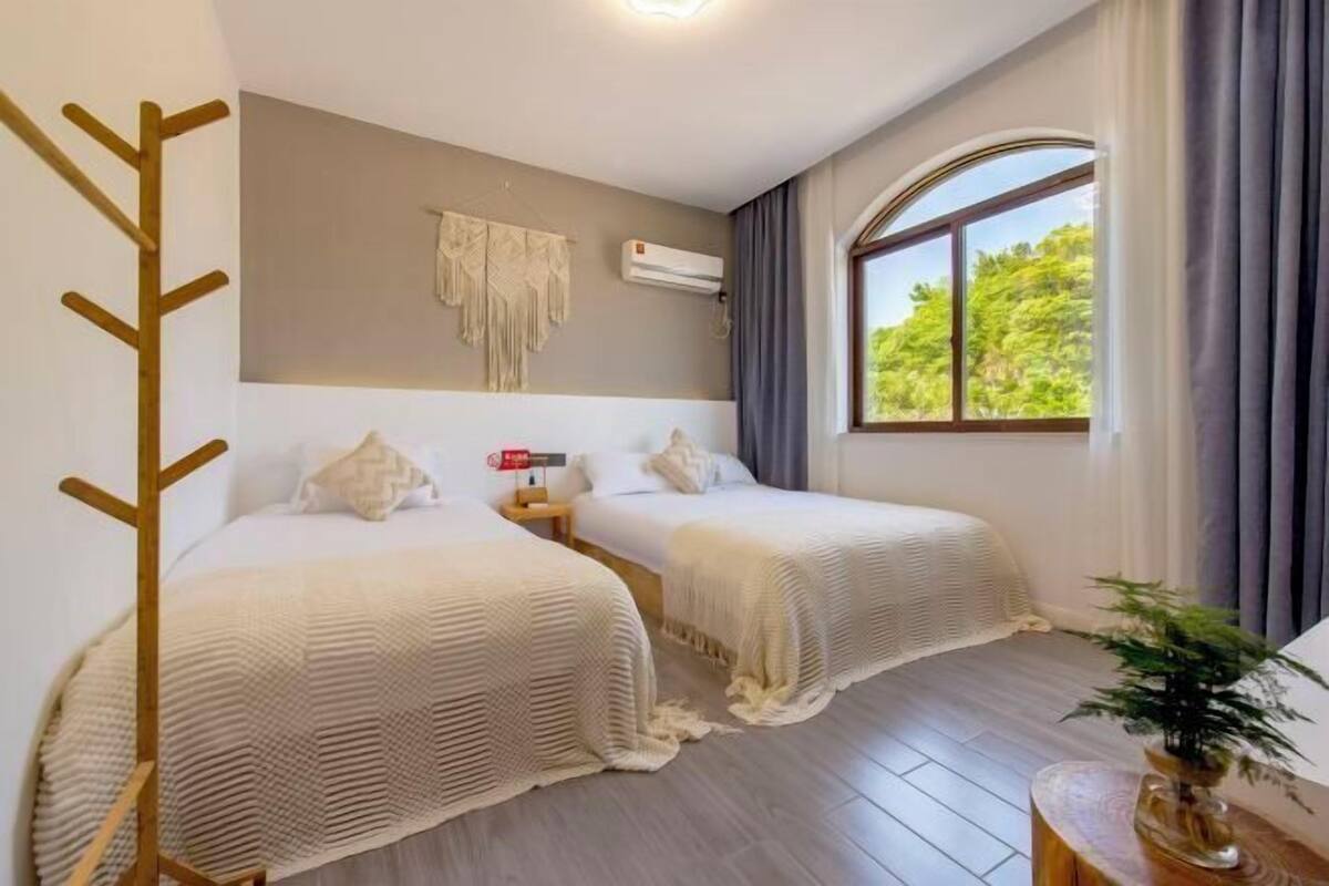 Ein Hotelzimmer mit zwei Betten, einem Holz-Kleiderständer, einer Klimaanlage, einem Fenster mit Vorhängen und einer Pflanze auf einem Beistelltisch.