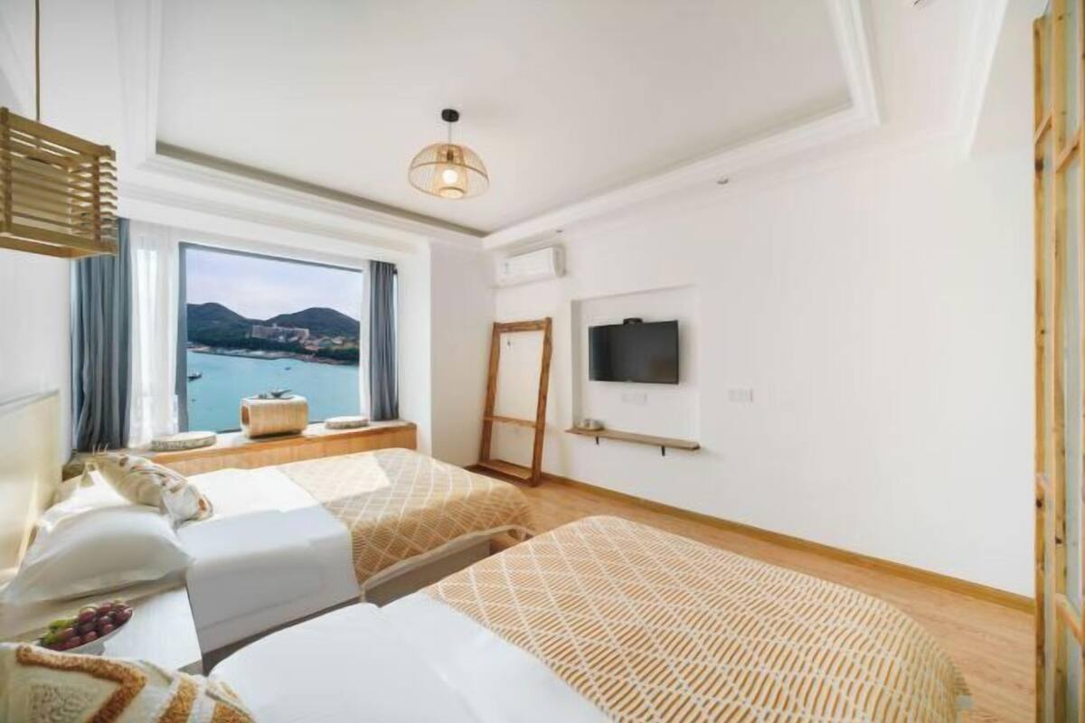 Ein Hotelzimmer mit zwei Betten, einem Fernseher, einem großen Fenster mit Meerblick und einer Deckenleuchte.