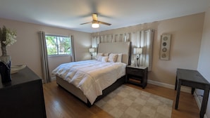 A bedroom with a bed, bedside tables, a ceiling fan, a window with curtains, and a small table with a lamp.