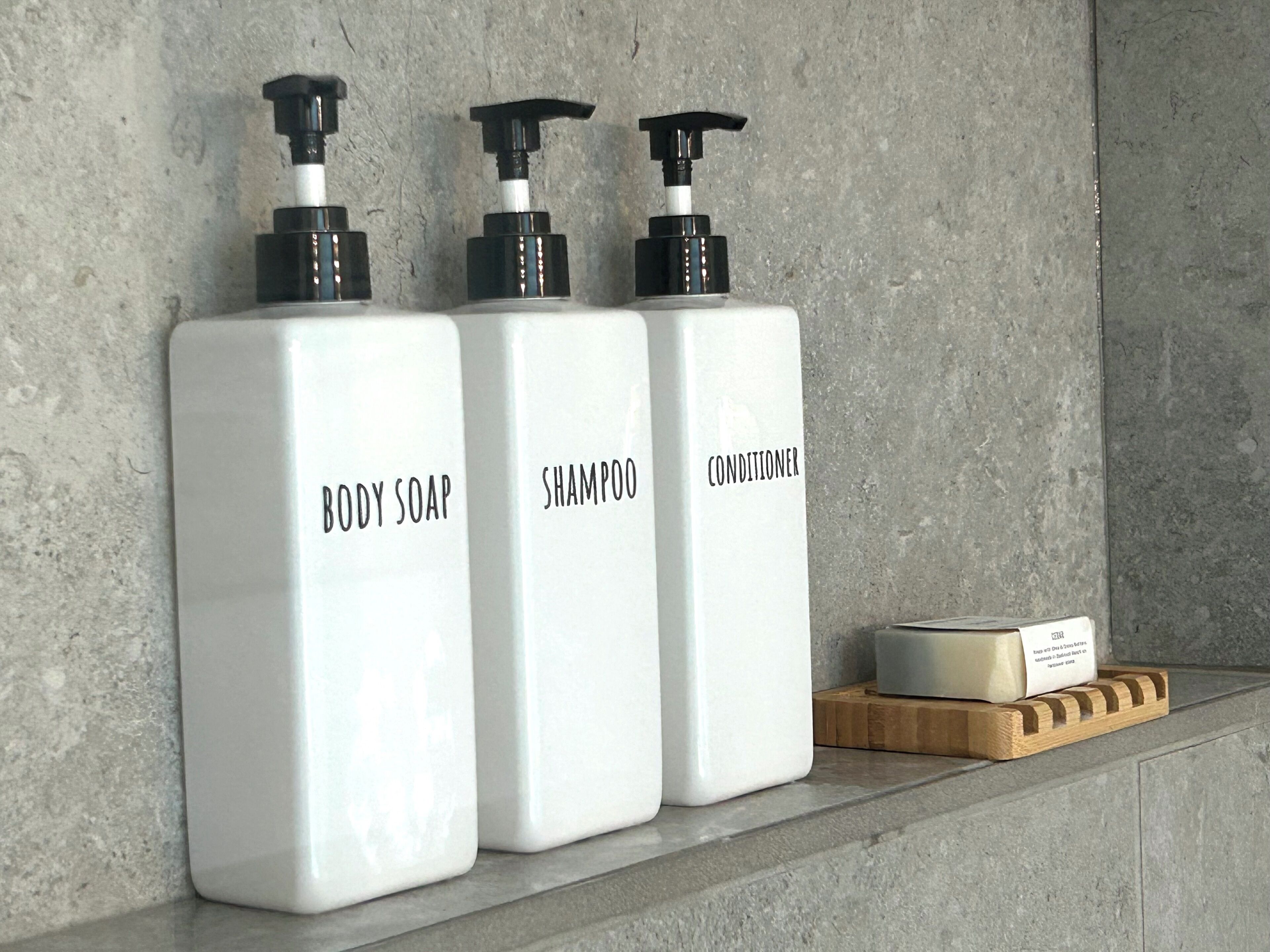Tres botellas blancas con etiquetas que indican jabón corporal, champú y acondicionador sobre una repisa contra una pared de hormigón.