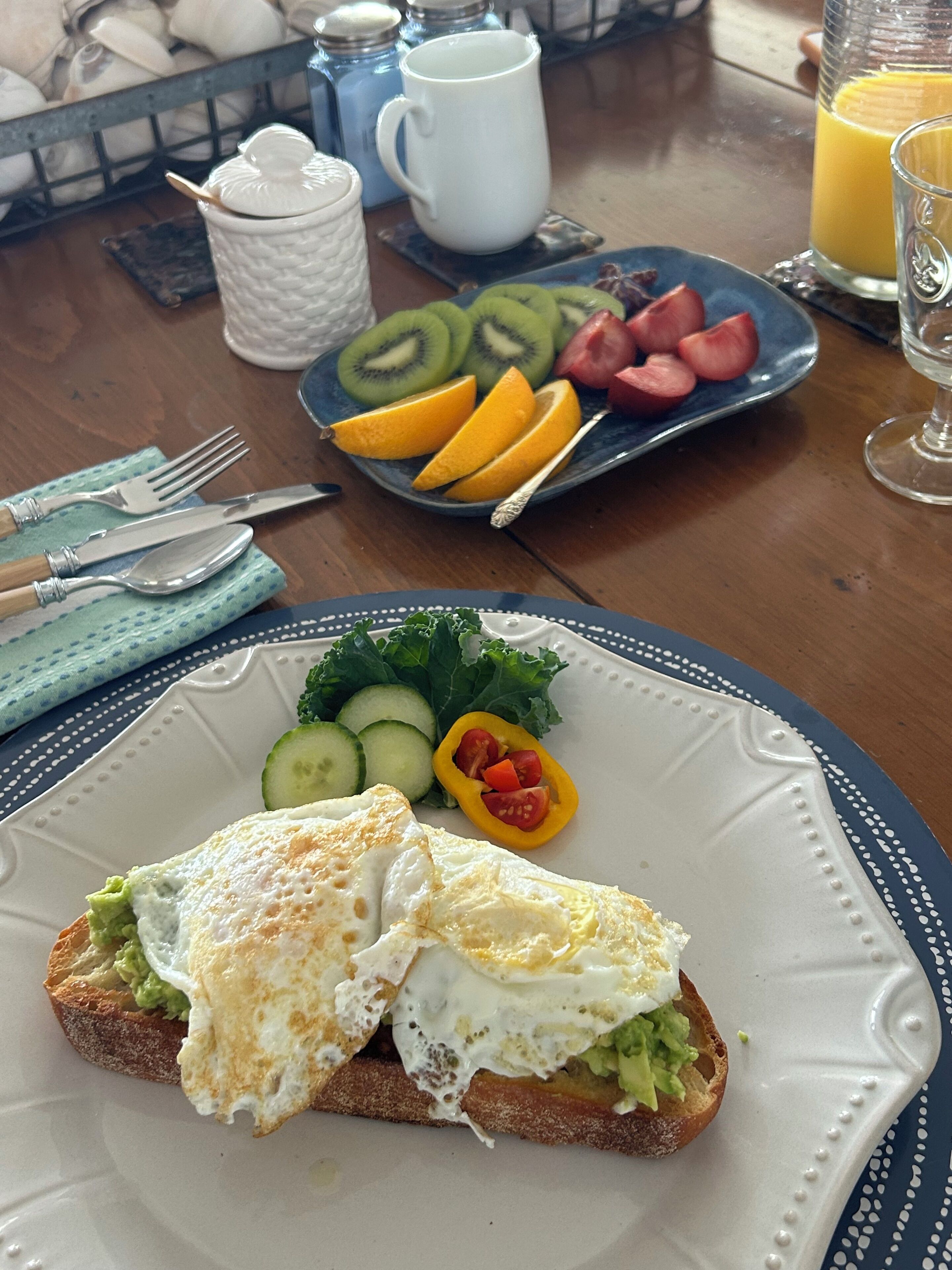 A breakfast setting with a plate of toast topped with avocado and egg, a side salad, and a plate of fruit including kiwi and orange slices.