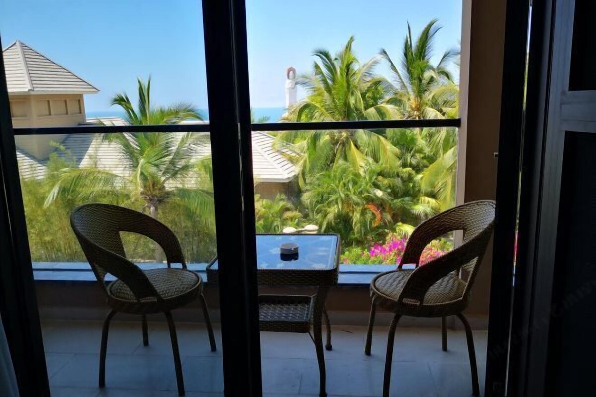 A balcony with wicker chairs and a small table, overlooking a garden with palm trees and a view of the ocean.