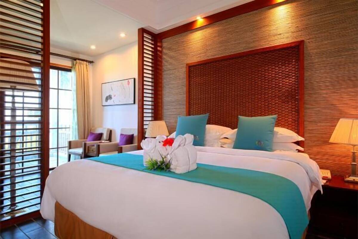 A hotel room with a large bed, a wooden headboard, a desk with a lamp, and a window with blinds.