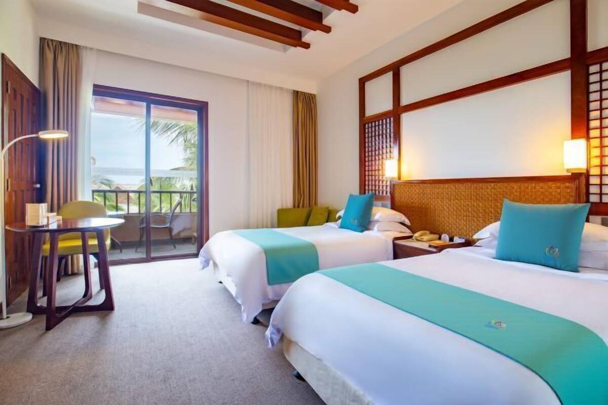 A hotel room with two beds, a balcony with a view, a desk with a chair, and a lamp.