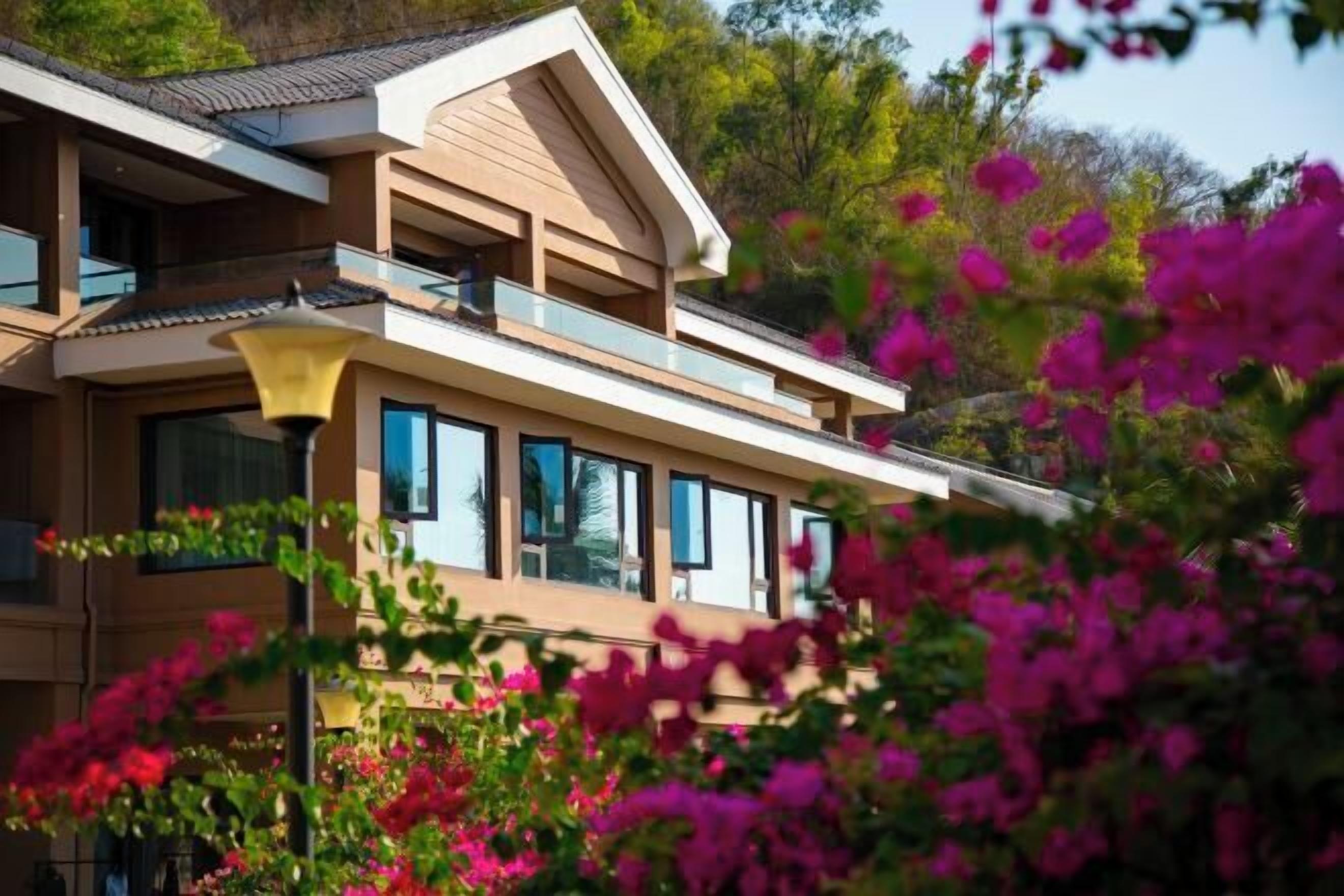 A modern two-story building with large windows and balconies, surrounded by lush greenery and vibrant flowers.
