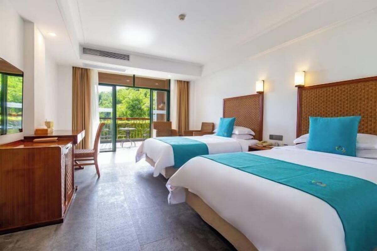 A hotel room with two beds, a desk, and a balcony with a view of greenery.