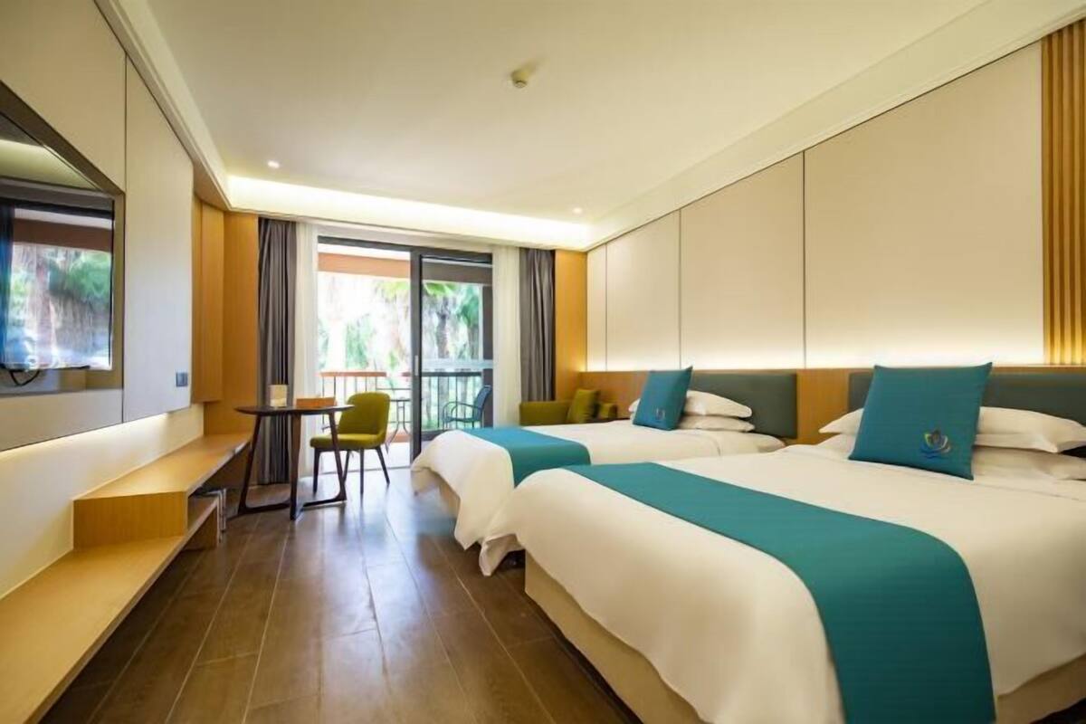 A hotel room with two beds, a desk, a chair, and a view of greenery.