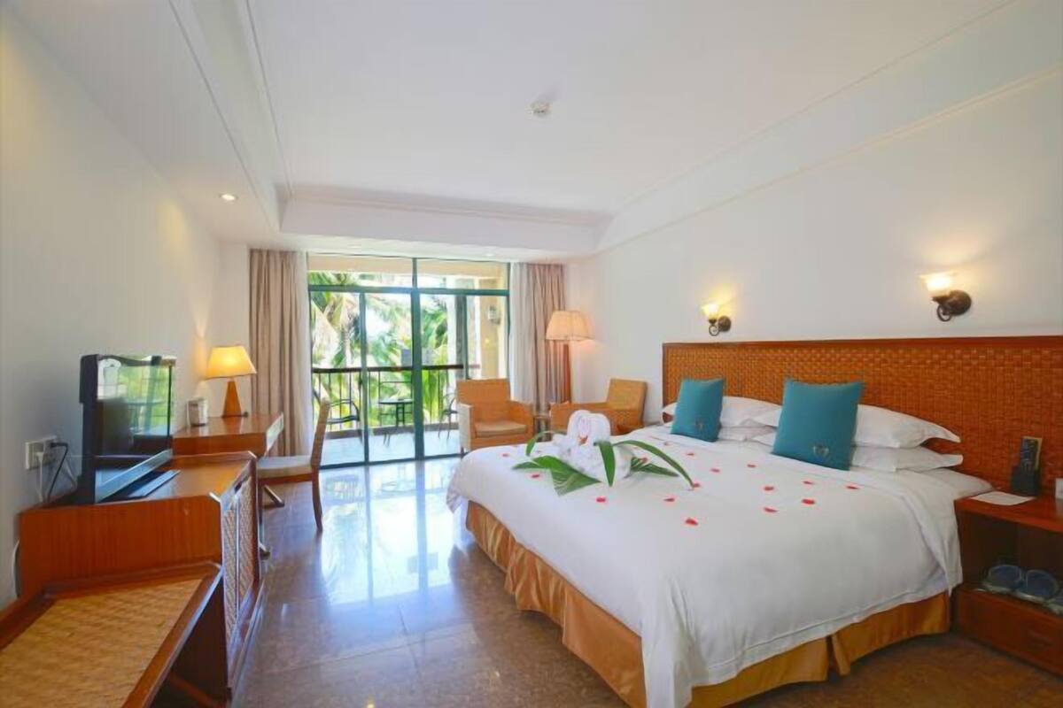 A hotel room with a large bed, a television, a desk, and a balcony with a view of greenery.