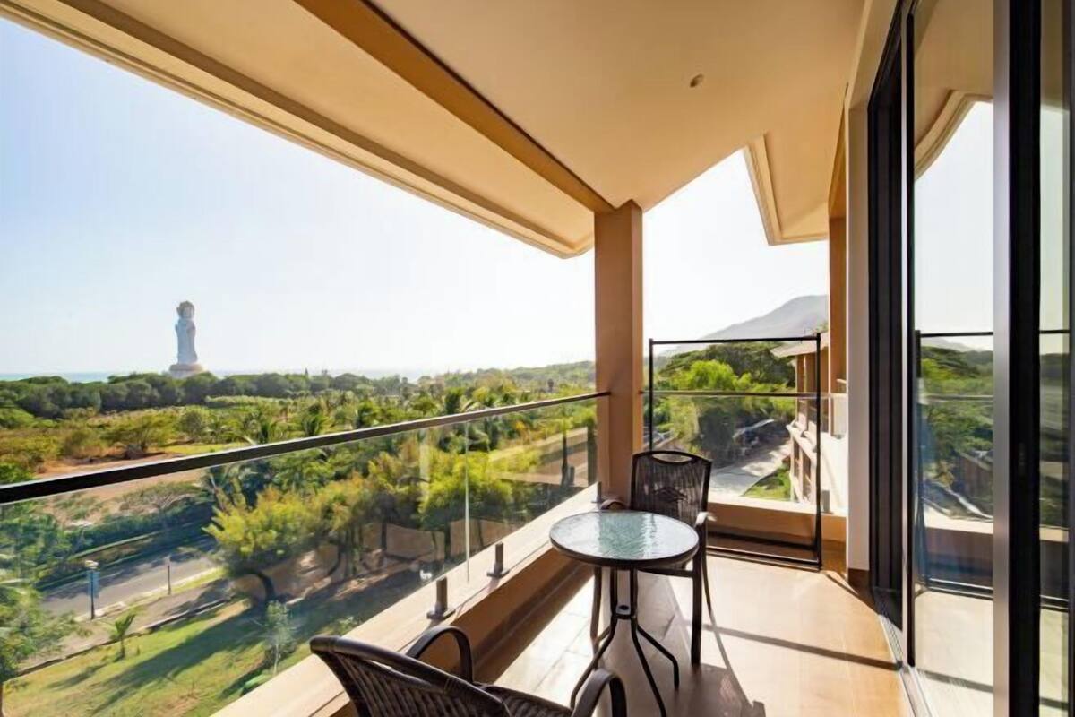 A balcony with a glass table and chairs, offering a view of greenery and a distant tower.