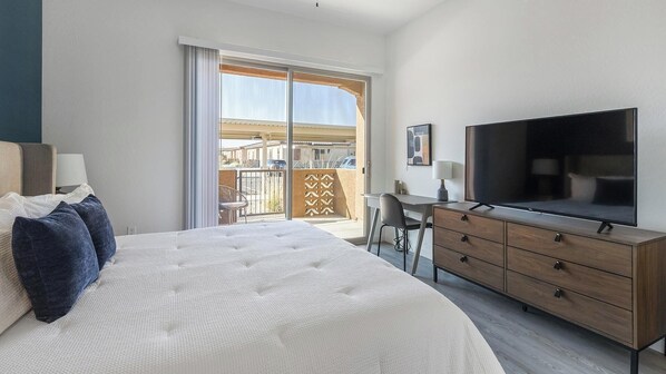 A hotel room with a large bed, a TV, a desk, and a balcony with a view of buildings.