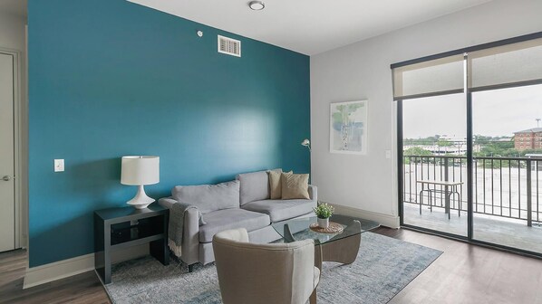 A modern living room with a teal accent wall, a grey sofa, a glass coffee table, and a view of the city through large sliding doors.