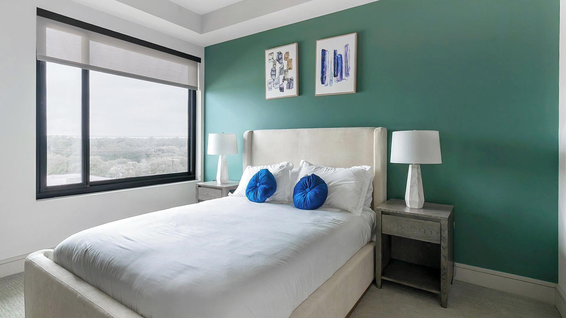 A hotel room with a large bed, two bedside tables with lamps, a window with a view, and two framed artworks on the wall.