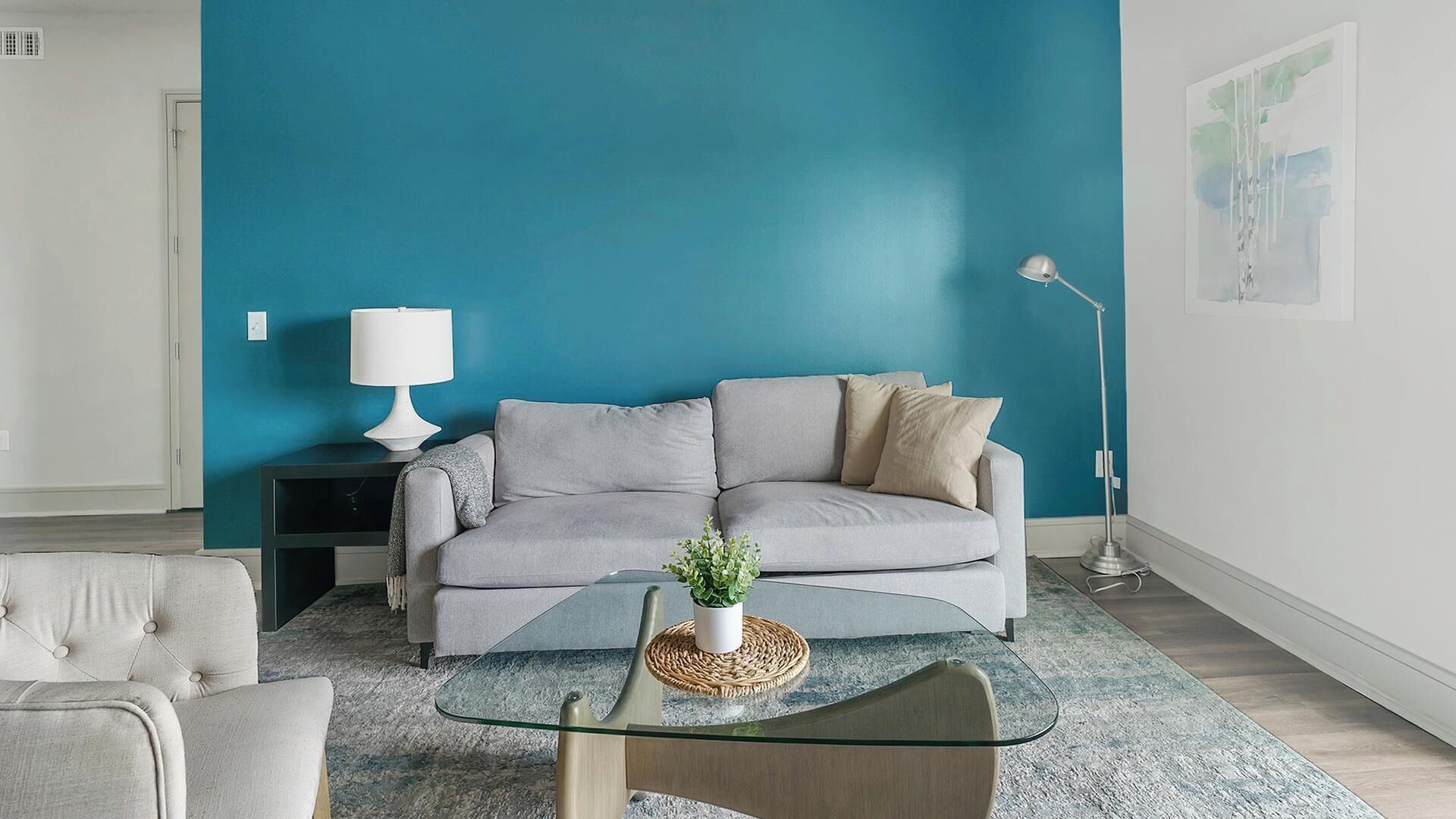 A living room with a teal wall, a grey sofa, a glass coffee table with a plant, and a floor lamp.