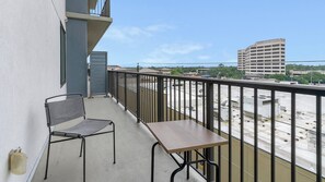 A balcony with a table and two chairs, a metal railing, and a view of a parking lot and buildings in the background.