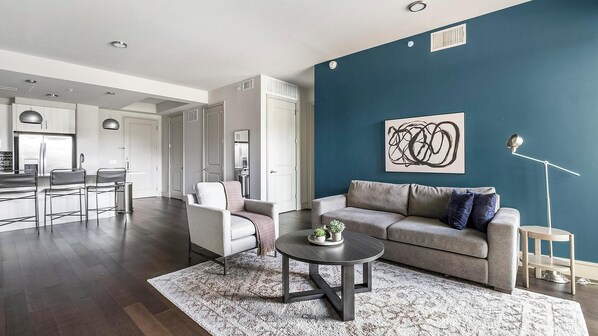 A modern living room with a dark wood floor, a grey sofa, a round coffee table, and a blue accent wall. - Landing | Stylish 2BD, Courtyard, Gym (Houston)