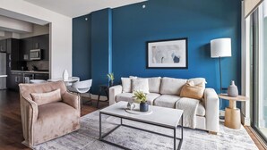 A modern living room with a blue accent wall, a sofa, a coffee table, and a minimalist kitchen in the background. - Landing | Modern 1BD, Pool, Gym (Houston)