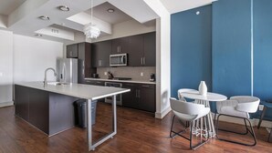 A modern kitchen with dark cabinets, a stainless steel refrigerator, a microwave, and a central island with a sink. There is a dining area with a round table and two chairs. - Landing | Modern 1BD, Pool, Gym (Houston)