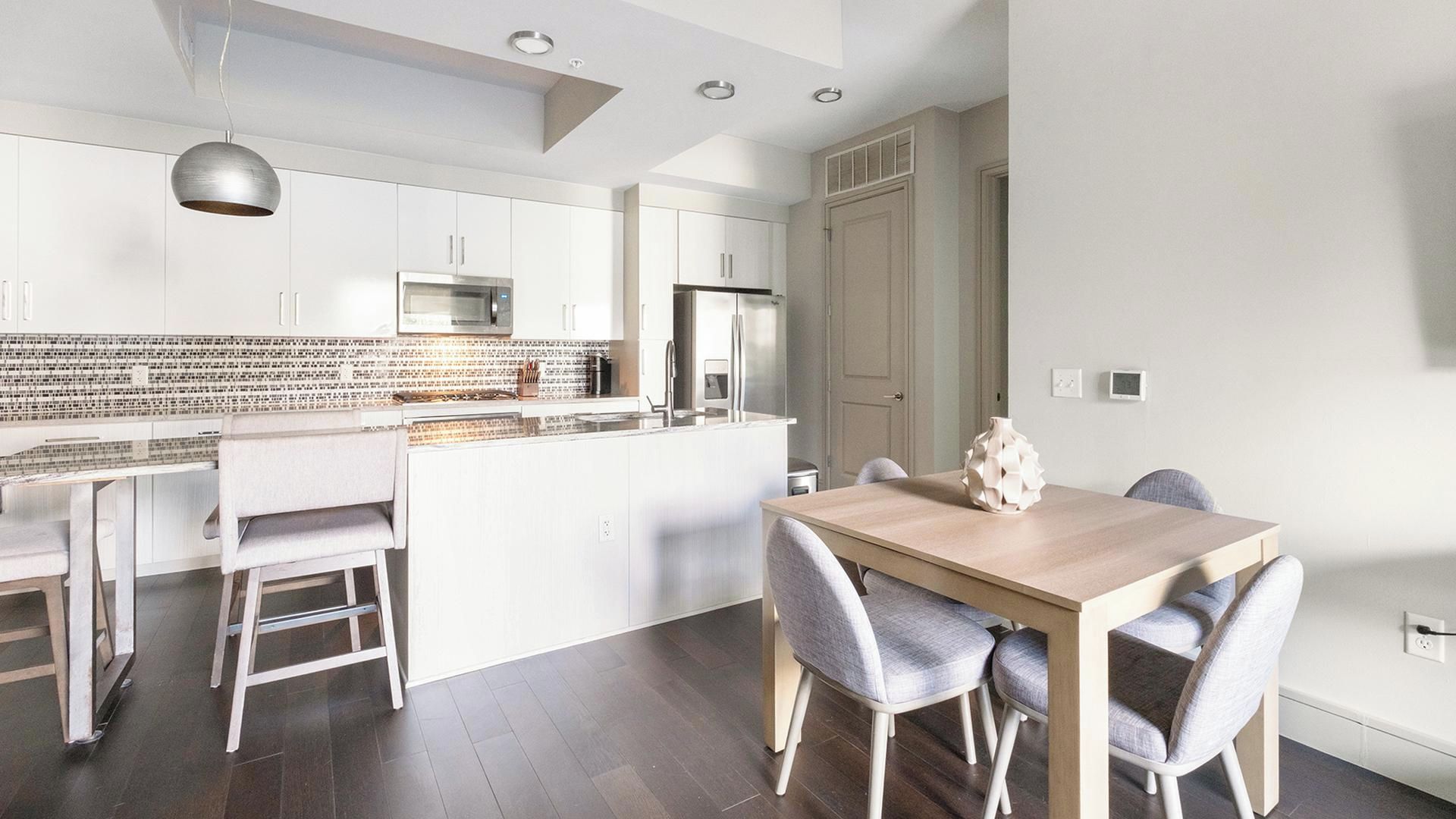 A modern kitchen with white cabinetry, a central island, and a dining area with a wooden table and grey upholstered chairs.