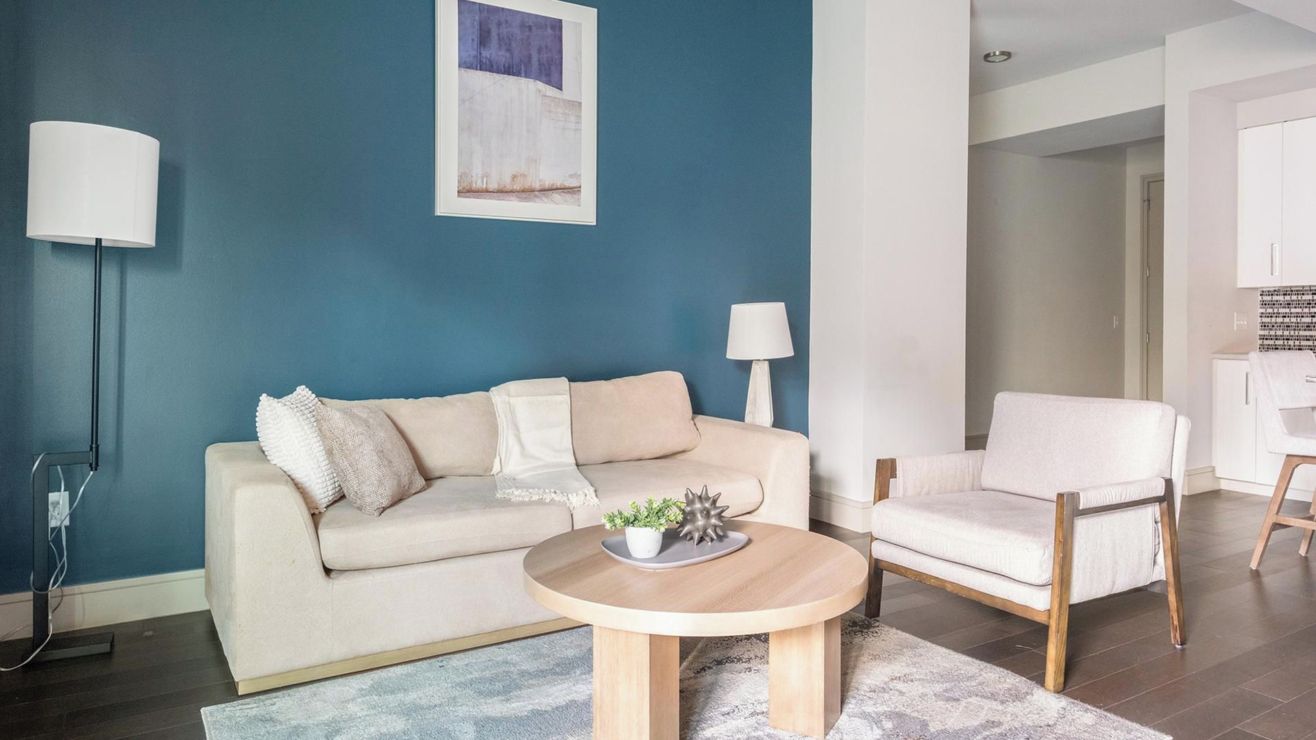 A living room with a beige sofa, a round wooden coffee table, and a white armchair. There is a painting on the wall and a lamp on a side table.