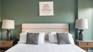 A hotel room with a bed, two bedside tables with lamps, a headboard, and a wall-mounted picture. - Landing | Beautiful 2BD, Pool, Gym (Houston)