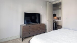 A bedroom with a bed, a dresser with a television on top, and an open door leading to a kitchen area.