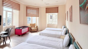 Une chambre d’hôtel avec trois lits, un parc pour enfants rouge et noir, un fauteuil beige et un bureau avec une chaise.