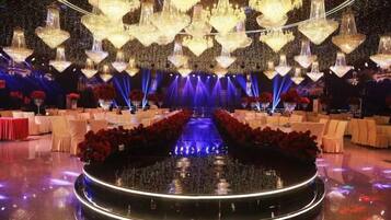 A formal event space with a central stage, multiple chandeliers, and rows of tables with chairs.
