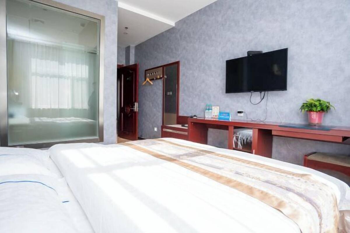 Hotellihuone, jossa on sänky, seinälle asennettu televisio, peilillä varustettu työpöytä ja ruukussa oleva kasvi.