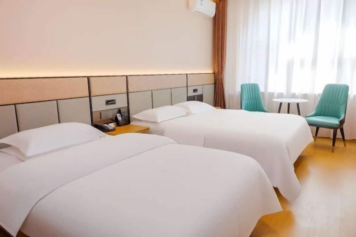 Hotellihuone, jossa on kaksi sänkyä, pieni pöytä, kaksi tuolia, yöpöytä ja ikkuna, jossa on verhot.