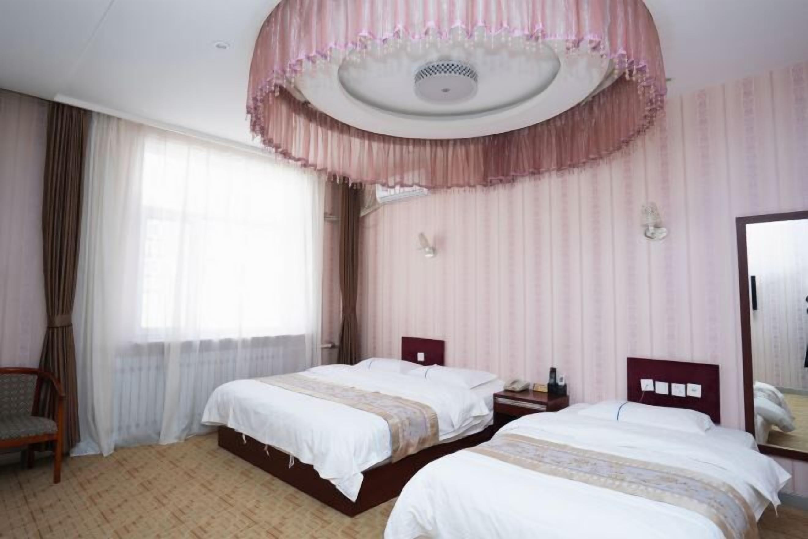 A hotel room with two beds, a wooden chair, a bedside table with a lamp, and a large mirror on the wall.